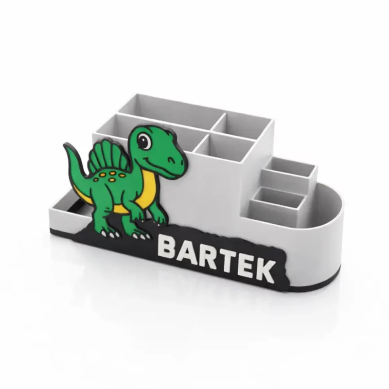 Biały przybornik szkolny z motywem uśmiechniętego zielonego spinozaura pokazany pod kątem. Układ przegródek ułatwiających organizację biurka dziecka.